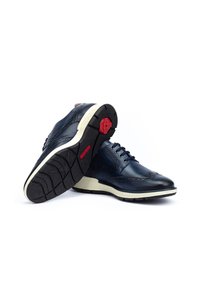 Chaussures en cuir bleu marine avec des détails en brogue, semelles intermédiaires blanches et semelles extérieures en caoutchouc noir texturé. Accents rouges sur le talon et la semelle.