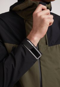 Giacca impermeabile nera e verde oliva con colletto alto, chiusura a zip e polsini regolabili, caratterizzata da planche in Velcro nero testurizzato e accenti con etichetta.