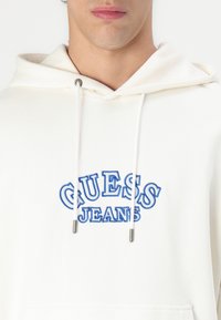 Sudadera blanca hecha de algodón. Presenta un gran logo bordado en azul "GUESS JEANS" en el frente, con cordones y puntas de metal plateado.