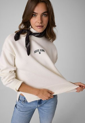 Frau trägt weißen Guess Jeans Pullover, schwarzen Schal um den Hals gebunden und blaue Jeans, hält den Pullover-Saum mit beiden Händen.