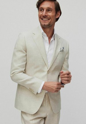 Lichtbeige linnen pak met een getailleerde pasvorm, enkelvoudig geknoopte blazer, wit overhemd en een gedessineerde pochet. Gladde textuur, minimale accenten.
