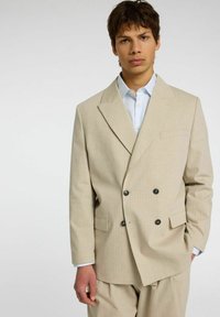 Giacca da abito beige doppiopetto con sottili righe verticali, rever strutturati e due tasche anteriori. Indossata sopra una camicia azzurra chiaro.