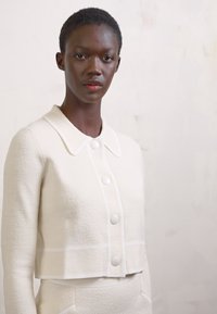 Chaqueta cortada en marfil con cuello, ribete blanco y botones redondos. Tejido texturizado, diseño ajustado, combinada con una parte inferior a juego.