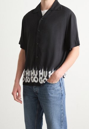 Homme portant une chemise noire à manches courtes avec un délavage blanc et un texte déformé à l'ourlet, associée à un jean bleu, main dans la poche.