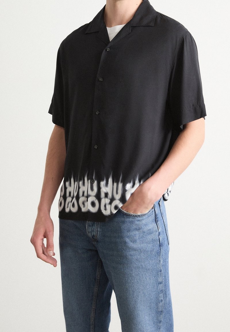 Homme portant une chemise noire à manches courtes avec un délavage blanc et un texte déformé à l'ourlet, associée à un jean bleu, main dans la poche.