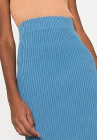 Zribovaná modrá sukně s vysokým pasem, vyrobená z měkké tkaniny. Materiál má vertikální pruhy a je kombinován s oranžovým zribovaným topem.