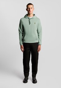 Felpa verde chiaro con cappuccio e tasca frontale, con un piccolo logo giallo sul petto, abbinata a pantaloni scuri e stivali neri.