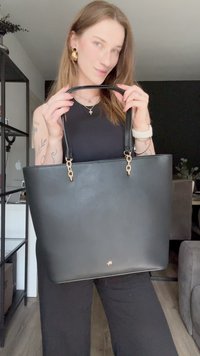 Bolsa tote de couro preta com alças duplas, detalhes em metal dourado e uma textura suave. Apresenta um design minimalista e abertura superior.