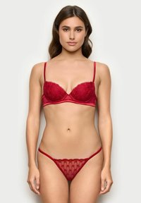 Conjunto de lencería roja que incluye un sujetador acolchado de encaje con tirantes ajustables y un tanga transparente a juego con borde en forma de ondulación y patrón floral.