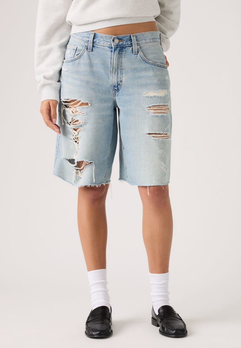 Φθαρμένα βερμούδα από τζιν σε ανοιχτό γαλάζιο χρώμα, φορεμένα με λευκές κάλτσες και μαύρα  μοκασίνια, εμφανίζονται σε ένα άτομο μπροστά σε απλό φόντο.