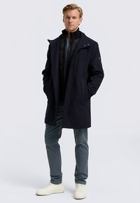 Tumši zilais pārvalks ar pogu un rāvējslēdzēja aizdari, valkāts virs melnas pūkaina vesta. Apģērbu papildina šauras pelēkas bikses un baltas sporta kurpes.