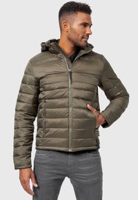 Chaqueta acolchada en verde oliva, con cierre frontal con cremallera, capucha, diseño acolchado y dos bolsillos laterales. Material suave y ligero.