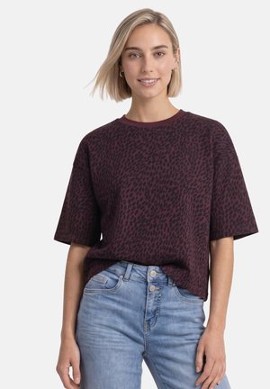 Ung kvinde med kort blond hår iført en mørkerød og sort mønstret oversized t-shirt og lyseblå højttaljede jeans.
