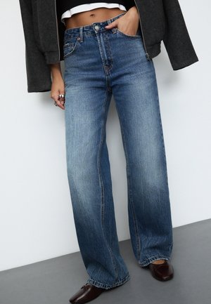 Personne portant un jean bleu taille haute à jambes larges, un haut blanc court, une veste foncée, et des chaussures pointues marron foncé sur un fond uni.