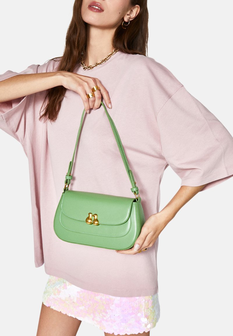 Grön läderhandväska med rund form, gyllene detaljer och axelrem, hålls av en person som bär en ljusrosa oversized skjorta.