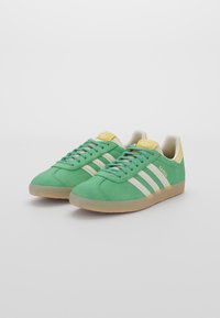 Sneaker in suede verde con strisce bianche, accenti gialli, design con lacci e suola in gomma. Logo "Gazelle" sul lato.