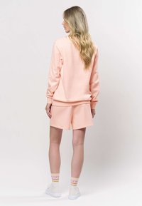 Světlounce růžová mikina a odpovídající kraťasy z měkké tkaniny. Mikina má dlouhé rukávy; kraťasy jsou ke kolenům. Bílé boty a vzorované ponožky.