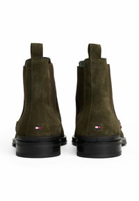 Un par de botas Chelsea de ante verde oliva con suelas negras, tiradores y pequeño logo de Tommy Hilfiger en la parte posterior.