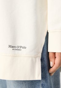 Gros plan sur un haut crème à manches longues avec le texte « Marc O'Polo EST. IN STOCKHOLM » au-dessus d'un jean en denim bleu et une main droite posée.