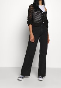 Kvinna iförd svarta vida jeans, svart stickad cardigan med vita knappar över ena axeln, vit ärmlös topp och svarta sneakers.