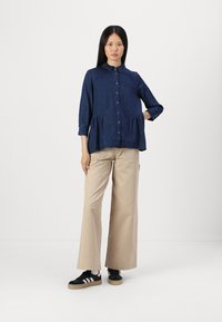 Dark blue denim button-up shirt with a peplum design and three-quarter sleeves, paired with beige wide-leg trousers and black sneakers.