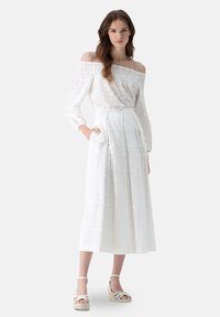 Weiße schulterfreie Bluse mit transparenter floralem Stickerei, kombiniert mit einem hochgeschnittenen, gefalteten weißen Rock mit Seitentaschen. Wedgesandalen.