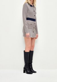 Abito in maglia beige chiaro con colletto e fascia in vita blu navy; caratterizzato da bottoni e una tasca frontale. Abbinato a stivali neri con tacco fino al ginocchio.