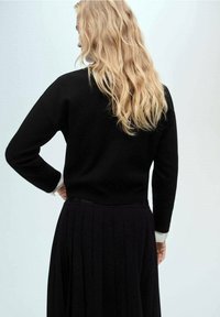 Femme blonde aux cheveux ondulés, vue de dos, portant un pull noir avec des poignets blancs et une jupe plissée noire.