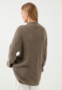 Brązowy sweter z dzianiny o luźnym kroju, z prążkowaną teksturą i wysokim kołnierzem. Posiada szerokie rękawy i nieco dłuższy tylny brzeg.