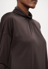 Maglia marrone a collo alto realizzata in un tessuto liscio e lucido; presenta un design plissettato all'scollatura e maniche a vestibilità normale.