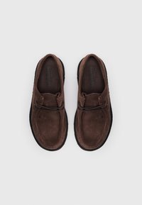 Sapatos de camurça castanhos com forma arredondada, solas de borracha pretas e atacadores decorativos. A palmilha apresenta texto em relevo.