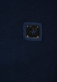 Marinblå stickad tröja med en strukturerad tyg. Har en fyrkantig svart etikett med pr Präglade "AM" initialer och fyra metall nitar.