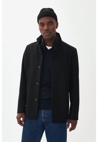 Cappotto nero con colletto strutturato, bottoni frontali e texture liscia; indossato sopra un maglione blu navy e jeans. Berretto a maglia nero.