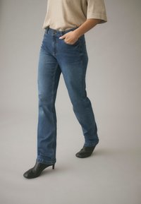 Person som bär blå jeans med raka ben, beige topp och svarta stövlar med klack, stående med ena handen i fickan mot en enfärgad bakgrund.