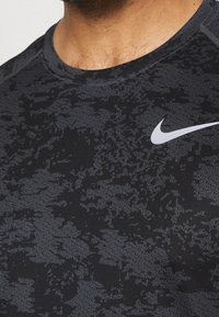Schwarzes Sportshirt mit einem strukturierten Muster in Dunkelgrau, mit einem weißen Nike-Logo auf der oberen linken Seite.