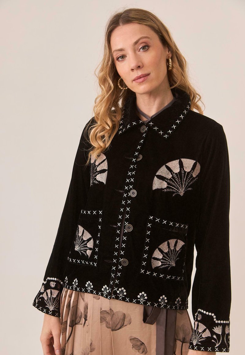 Mujer con largo cabello rubio ondulado lleva una chaqueta negra bordada con patrones florales sobre una falda beige con estampados florales oscuros.