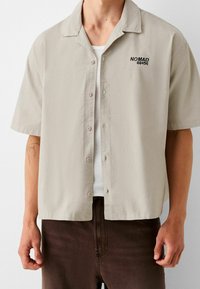 Camisa de botones de manga corta beige con cuello, que presenta texto negro bordado "NOMAD 00456" y una textura de tela suave. Llevada sobre una camiseta sin mangas blanca.