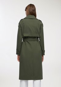 Oliwkowy trencz z paskiem w talii, z klasycznym kołnierzem, naramiennikami i długimi rękawami. Gładka tekstura materiału.