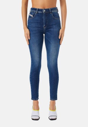 Personne portant un jean skinny taille haute bleu foncé avec un effet délavé sur les cuisses et des chaussettes blanches à bout carré avec un liseré jaune sur un fond blanc.
