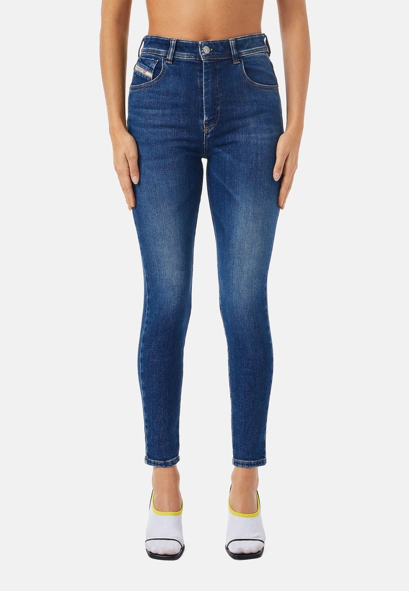 Personne portant un jean skinny taille haute bleu foncé avec un effet délavé sur les cuisses et des chaussettes blanches à bout carré avec un liseré jaune sur un fond blanc.