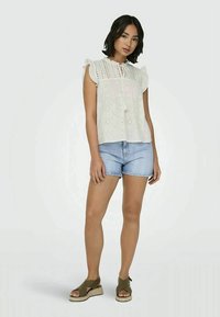 Blusa branca sem mangas com detalhes de ilhós, ombros com babados suaves, combinada com calções de ganga azul claro e sandálias verdes.