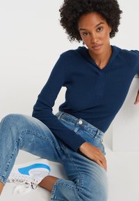 Femme aux cheveux bouclés portant un pull côtelé bleu foncé, un jean en denim bleu clair et des baskets blanches avec des accents bleus, assise de manière décontractée.
