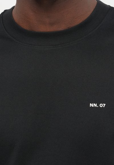 Prim-plan al unei persoane care poartă un tricou negru cu guler rotund, având inscripționat în alb textul „NN. 07” pe piept.