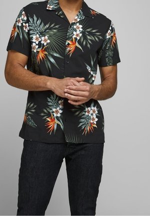 Hombre con camisa negra de manga corta abotonada con estampado tropical de flores blancas y naranjas y jeans oscuros, con las manos entrelazadas.
