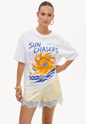 Femme portant un T-shirt blanc oversize avec un motif coloré de soleil et de vagues et le texte "Sun Chasers,", associé à un short jaune clair orné de dentelle.