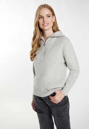 Donna con lunghi capelli biondi che indossa un maglione a maglia grigio chiaro con mezza cerniera e jeans scuri, sorridente e che guarda di lato.