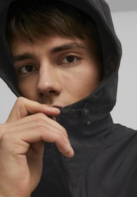 Svart vanntett jakke med høy krage, glidelås lukking og justerbar snorlukking. Glatt tekstur og tettsittende design. Hånd nær kragen.
