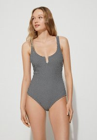 Černobílý kostkovaný jednodílný plavky s hlubokým výstřihem, širokými ramínky a vysokým vykrojením po stranách. Obsahuje detail klíčové díry vpředu.
