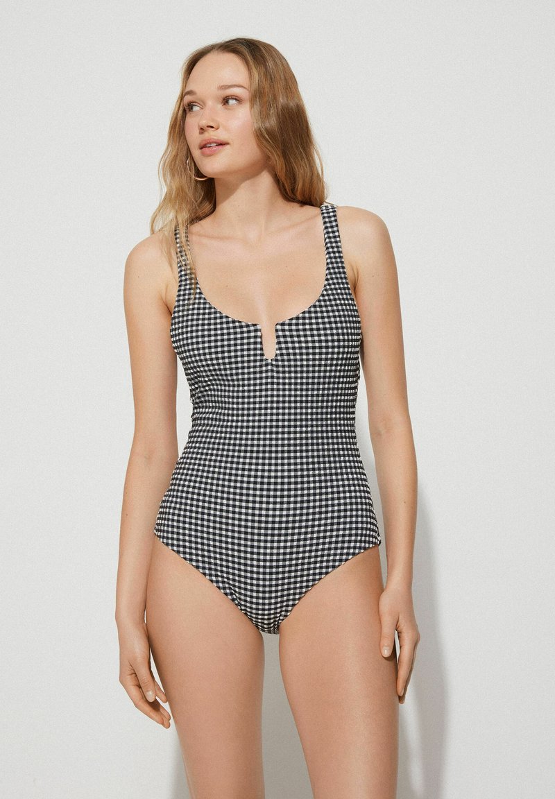 Černobílý kostkovaný jednodílný plavky s hlubokým výstřihem, širokými ramínky a vysokým vykrojením po stranách. Obsahuje detail klíčové díry vpředu.