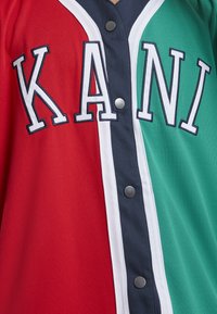 Punane ja roheline pesapallimängija särk musta nööpidele rihmaga ning suurete valgete kontuurjuhtmetega "KANI" rinnal.